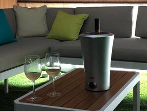 Ein modernes Wohnzimmer mit einem grauen Sofa, grünen und türkisfarbenen Kissen und einem weißen Couchtisch. Auf dem Tisch stehen zwei Weingläser und ein silbernes Wasserkocher.