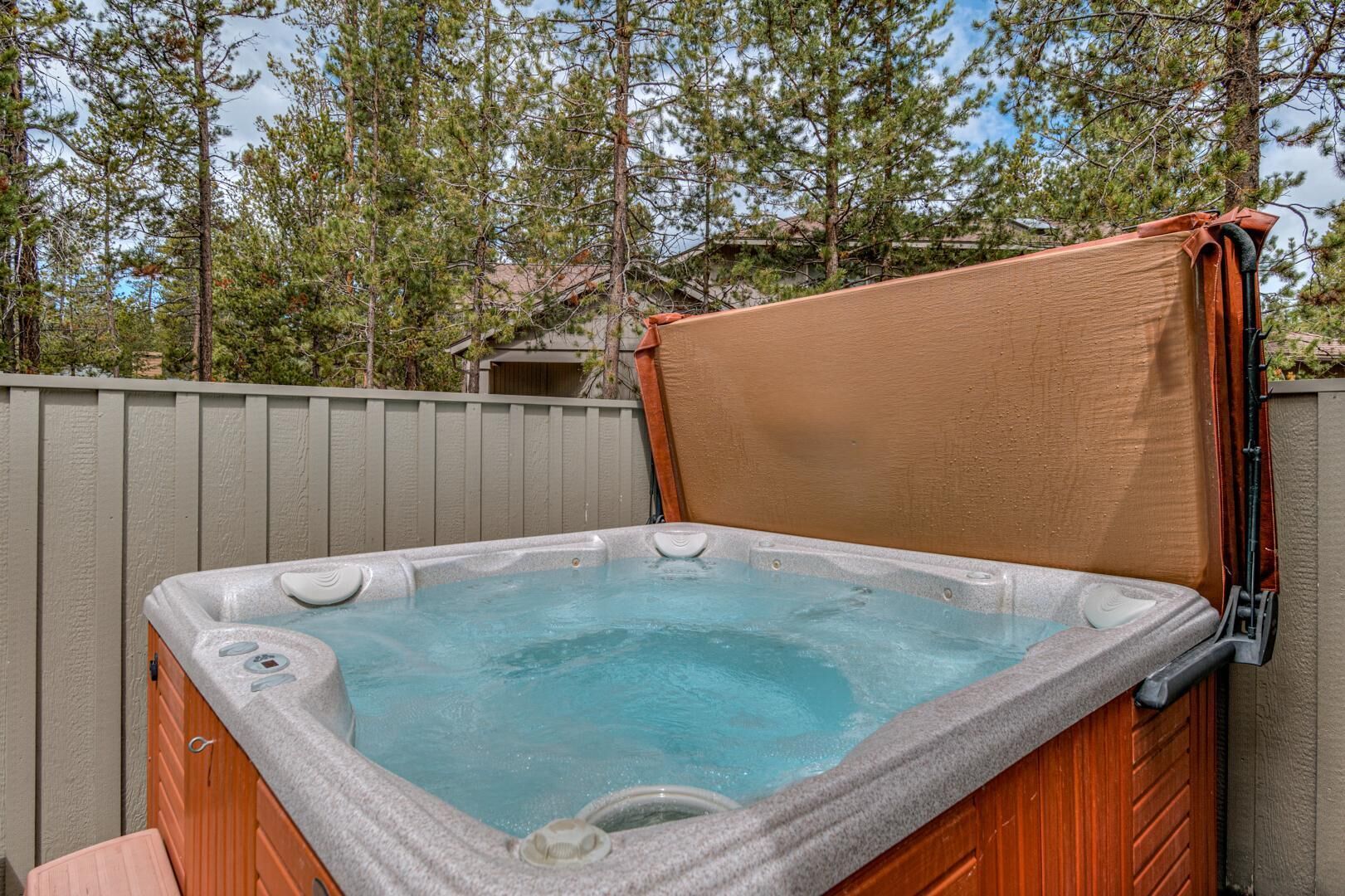 A hot tub with a wooden exterior and a beige cover, surrounded by a wooden fence and trees.