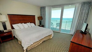 A hotel room with a large bed, a desk, a chair, a TV, and a balcony with a view of the ocean.