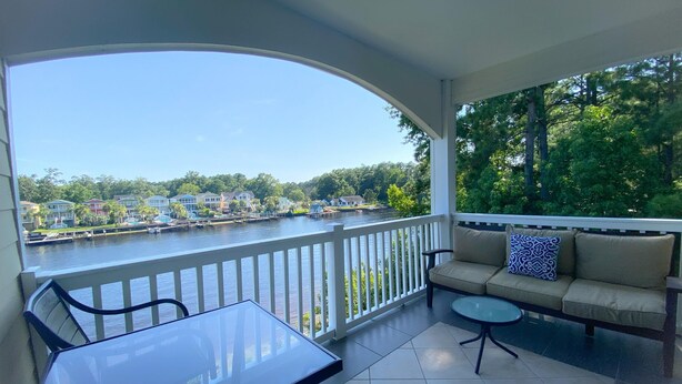 Een balkon met een glazen tafel en stoelen, een bank met kussens, en uitzicht op een rivier met huizen en bomen.