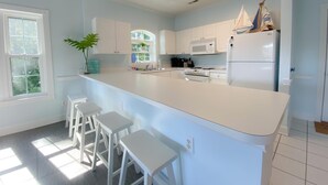 Een moderne keuken met witte kastjes, een centraal kookeiland met krukken, een magnetron, een koelkast en een raam met uitzicht op groen.