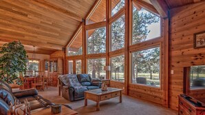 A spacious log cabin interior with large windows, a leather sofa, a dining area, and a fireplace. - Wood Duck Drive 55905 (Bend)