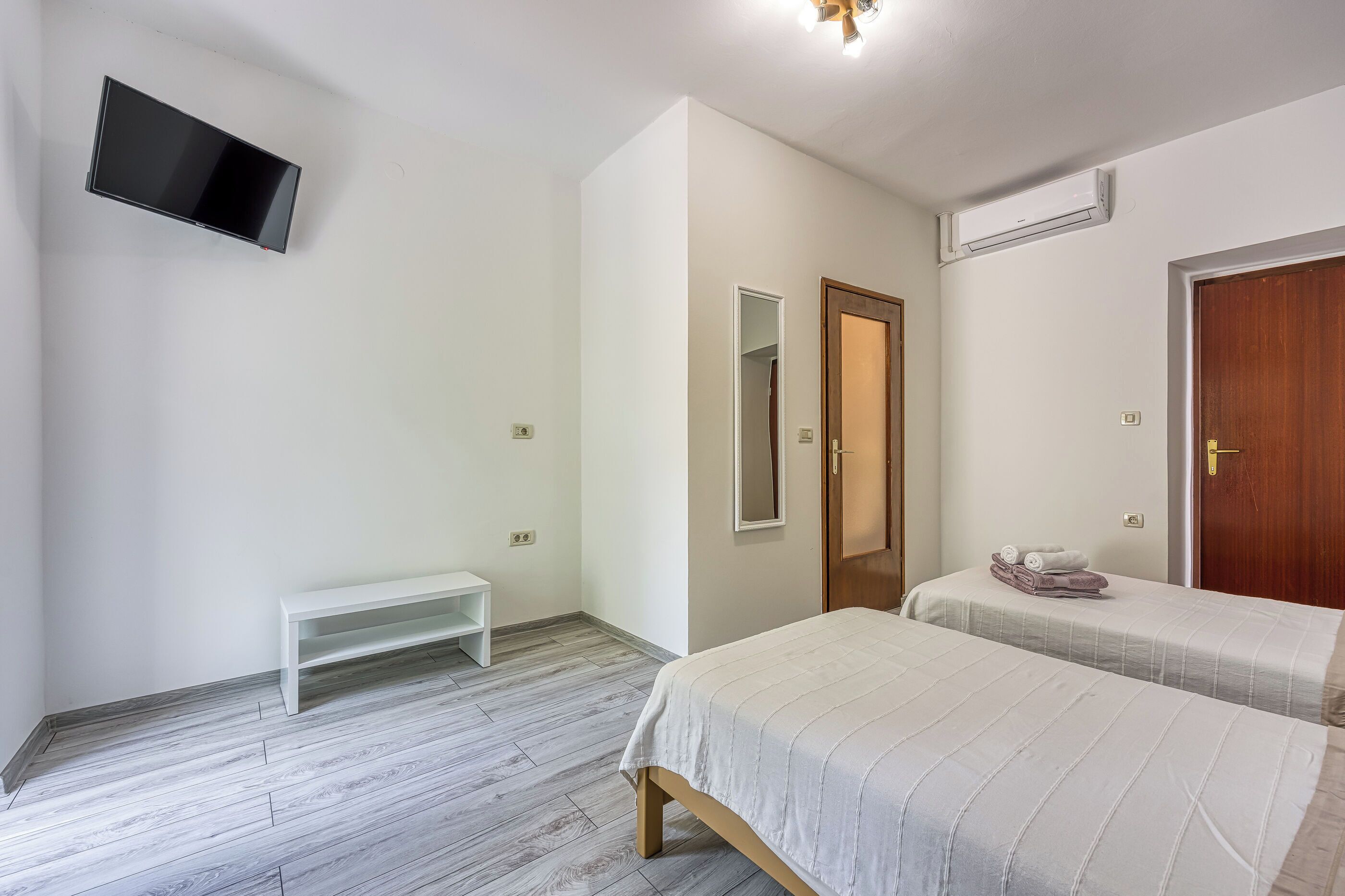 A bedroom with two beds, a TV, a bench, a mirror, and an air conditioning unit.