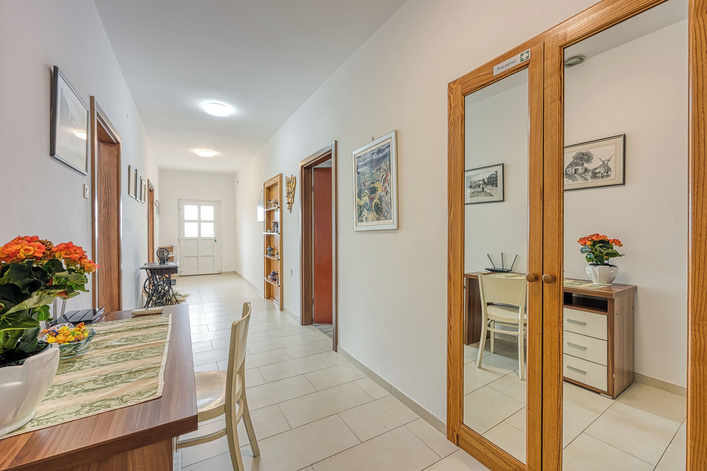 A hallway with a wooden desk, chairs, and a mirror. There are framed pictures on the walls and a window at the end of the hallway.