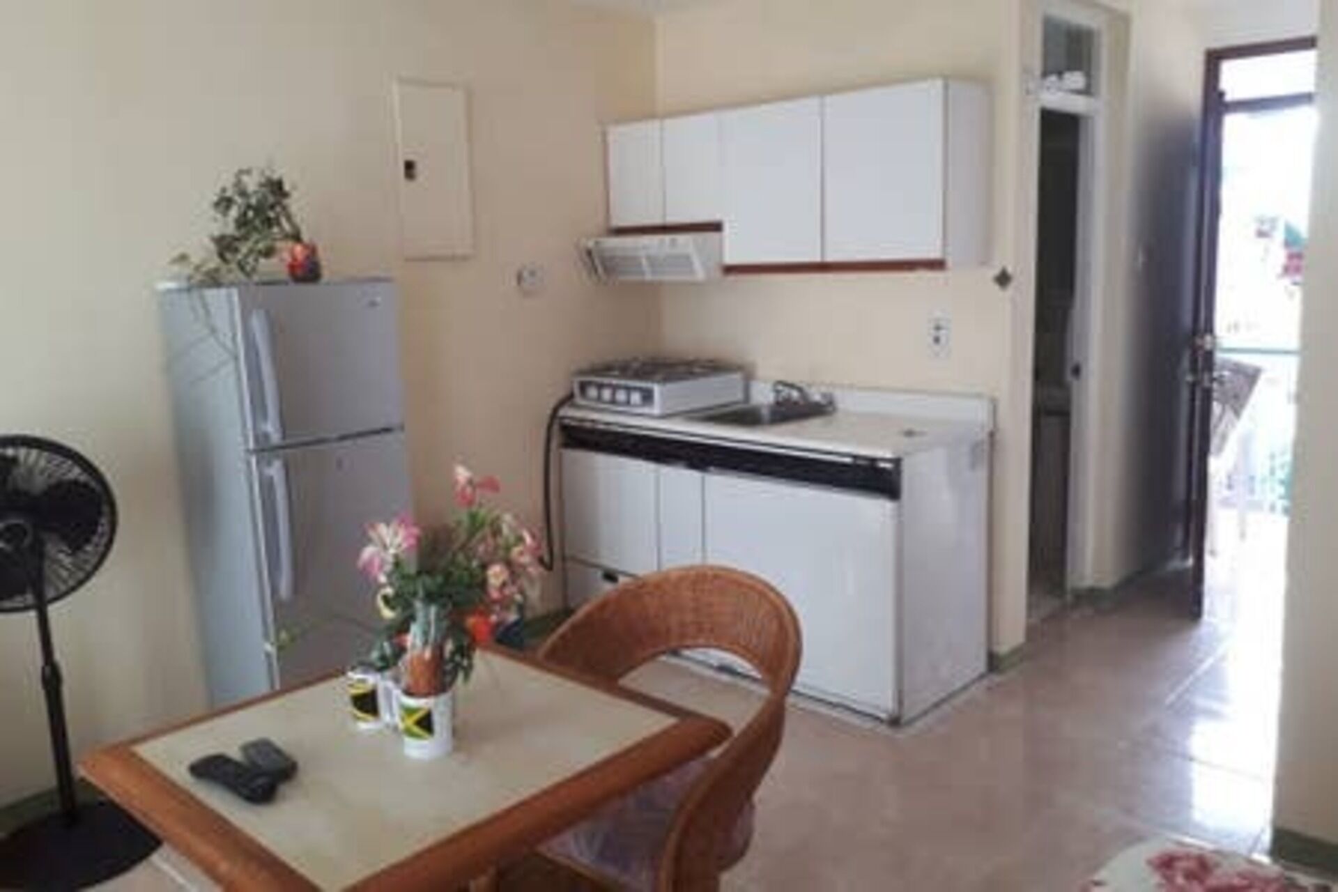 A compact kitchen with white cabinets, a small refrigerator, and a stove. There is a dining area with a table and chairs, and a fan.