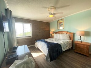 A bedroom with a bed, bedside lamps, a ceiling fan, a window with blinds, a desk with a computer, and a TV. - Ocean Forest Villas by Palmetto Vacations (Myrtle Beach)