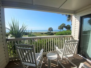 Een balkon met witte stoelen en een kleine tafel, uitkijkend op een strand met palmbomen en de oceaan.