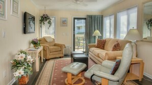 A cozy living room with a beige sofa, a chair, a small table, and a floor lamp. There is a ceiling fan, a window with blinds, and a door leading outside. - Ocean Forest Villas by Palmetto Vacations (Myrtle Beach)