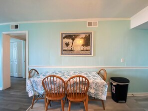A dining area with a table set for four, wooden chairs, a framed picture on the wall, and a trash can to the side. - Ocean Forest Villas by Palmetto Vacations (Myrtle Beach)