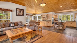 A spacious living room with a wooden ceiling, a fireplace, a dining area, and a kitchen with wooden cabinets and a stone countertop.