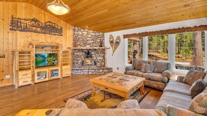 A cozy cabin interior with a stone fireplace, wooden furniture, and a large window offering a view of the forest.