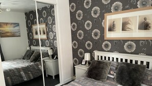 A bedroom with a mirrored closet door, a bed with a patterned bedspread, a nightstand with a lamp, and a wall with framed pictures.