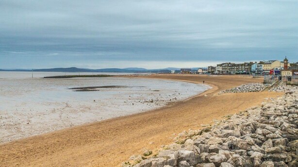 A coastal beach with a rocky shoreline, a sandy beach, and a row of buildings in the background.
