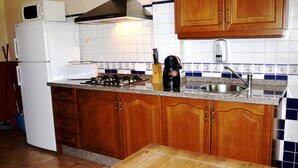 Egy konyha, melyben fa szekrények, egy fehér hűtőszekrény, egy tűzhely és egy mosogató található.