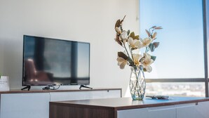 Une chambre d’hôtel moderne avec un téléviseur à écran plat posé sur un bureau en bois, un vase avec des fleurs et une grande fenêtre donnant sur la ville.