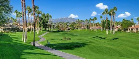 Un campo de golf con un camino despejado, palmeras y una montaña al fondo.