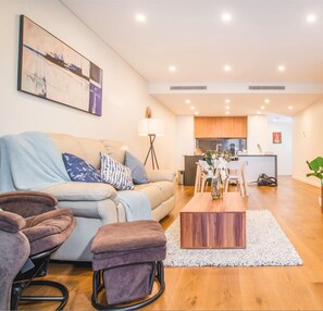A modern living room with a sofa, ottoman, and coffee table, a dining area with chairs, a kitchen in the background, and a painting on the wall.