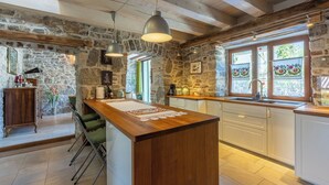 A rustic kitchen with stone walls, wooden beams, a central island, and a large window with decorative curtains. - House Carpe Diem (Jurdani)
