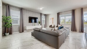 A modern living room with a grey sectional sofa, a flat-screen TV mounted on the wall, a potted plant, and a view of the outdoors through large windows. - 1596 MC - Enchanted 6BR Villa: Pool, Magic for 14 (Davenport)