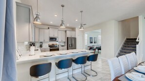 A modern kitchen with a central island, bar stools, and a dining area with a table and chairs. - 1596 MC - Enchanted 6BR Villa: Pool, Magic for 14 (Davenport)