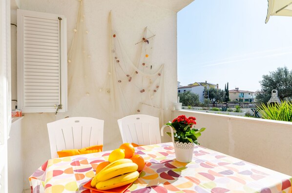 A balcony with a table set for two, featuring a colorful tablecloth, a fruit bowl with bananas and oranges, and a vase with flowers.