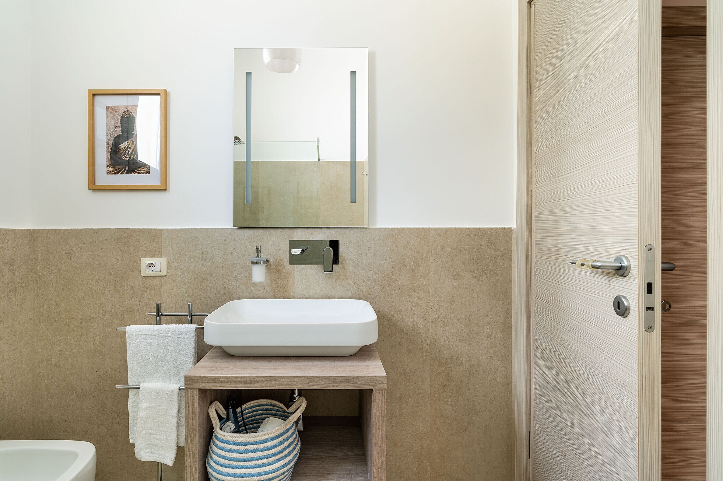 A modern bathroom with a white sink, a towel rack, a framed picture, and a wooden door.