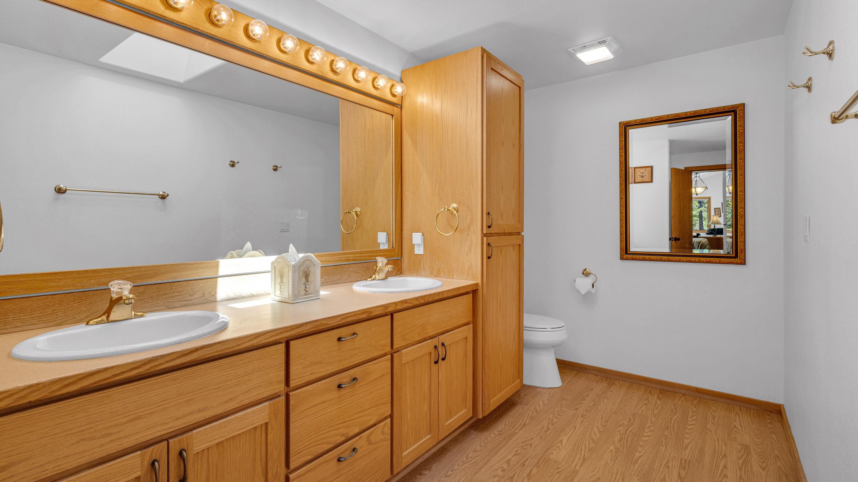 Une salle de bain avec des meubles en bois, deux lavabos, un miroir avec éclairage intégré, des toilettes et un miroir encadré accroché au mur.