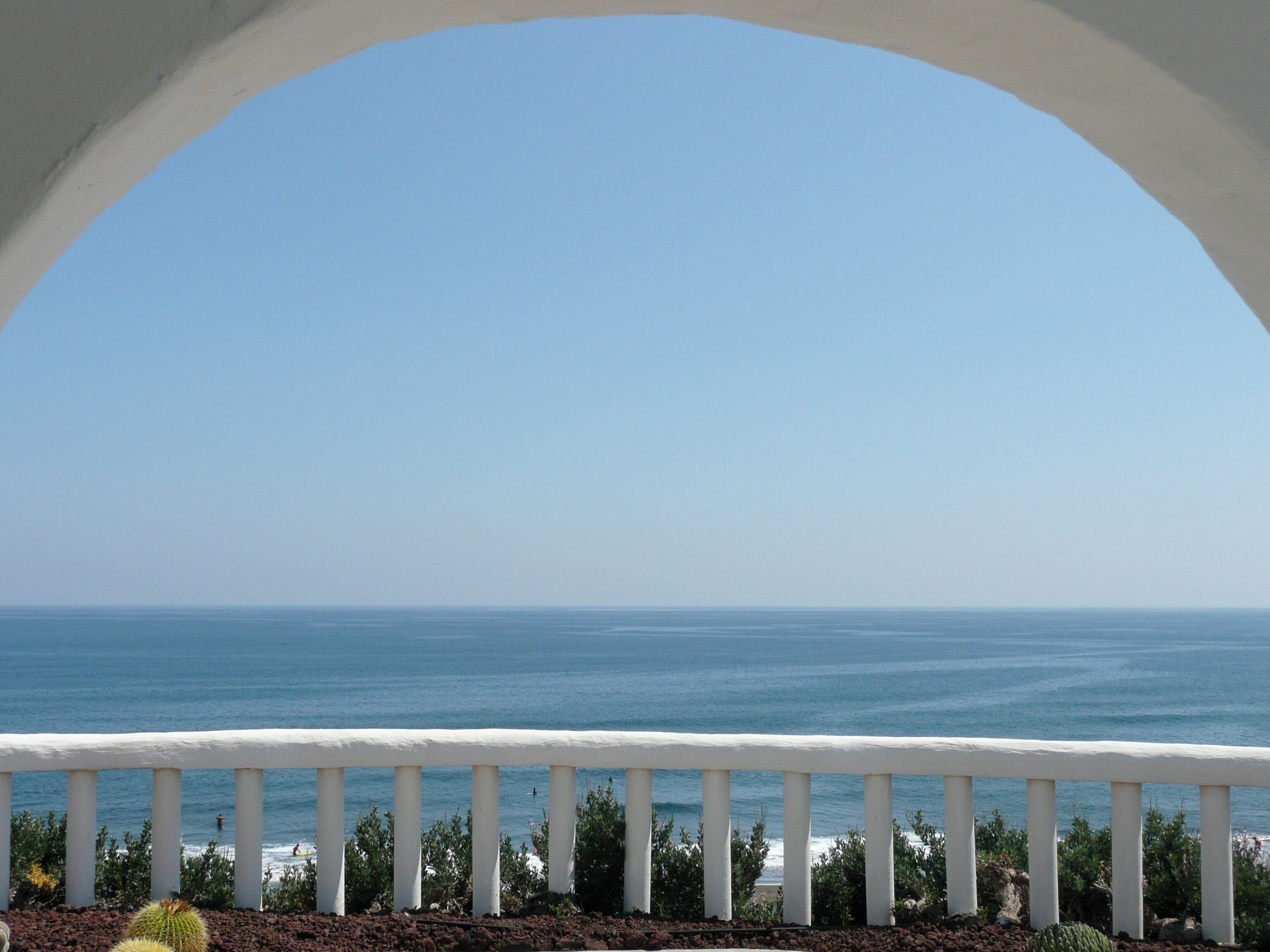 A coastal view through an arched window with a white railing, overlooking the sea.