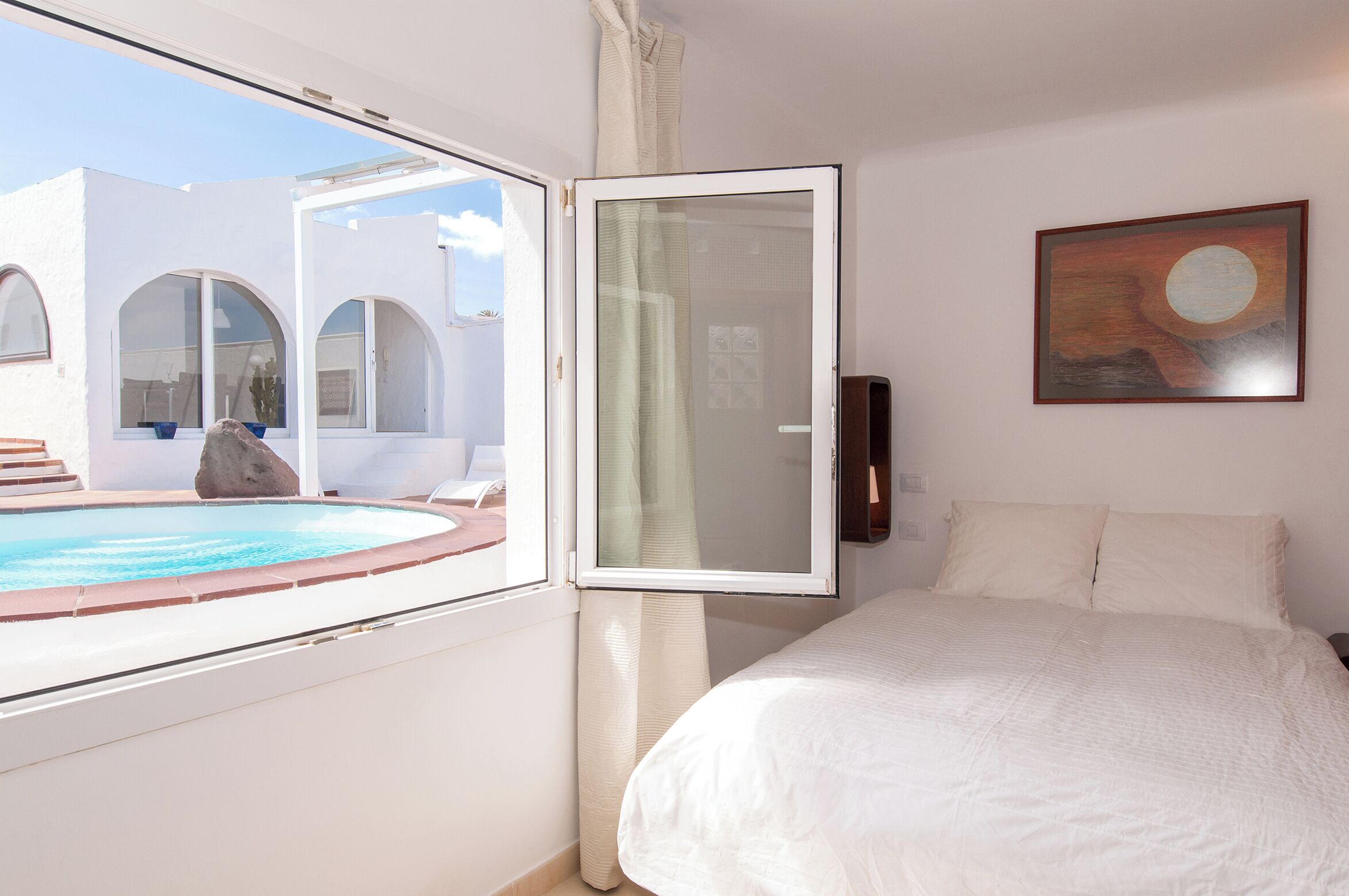 A bedroom with a bed, a painting, a view of a pool, and a white wall.