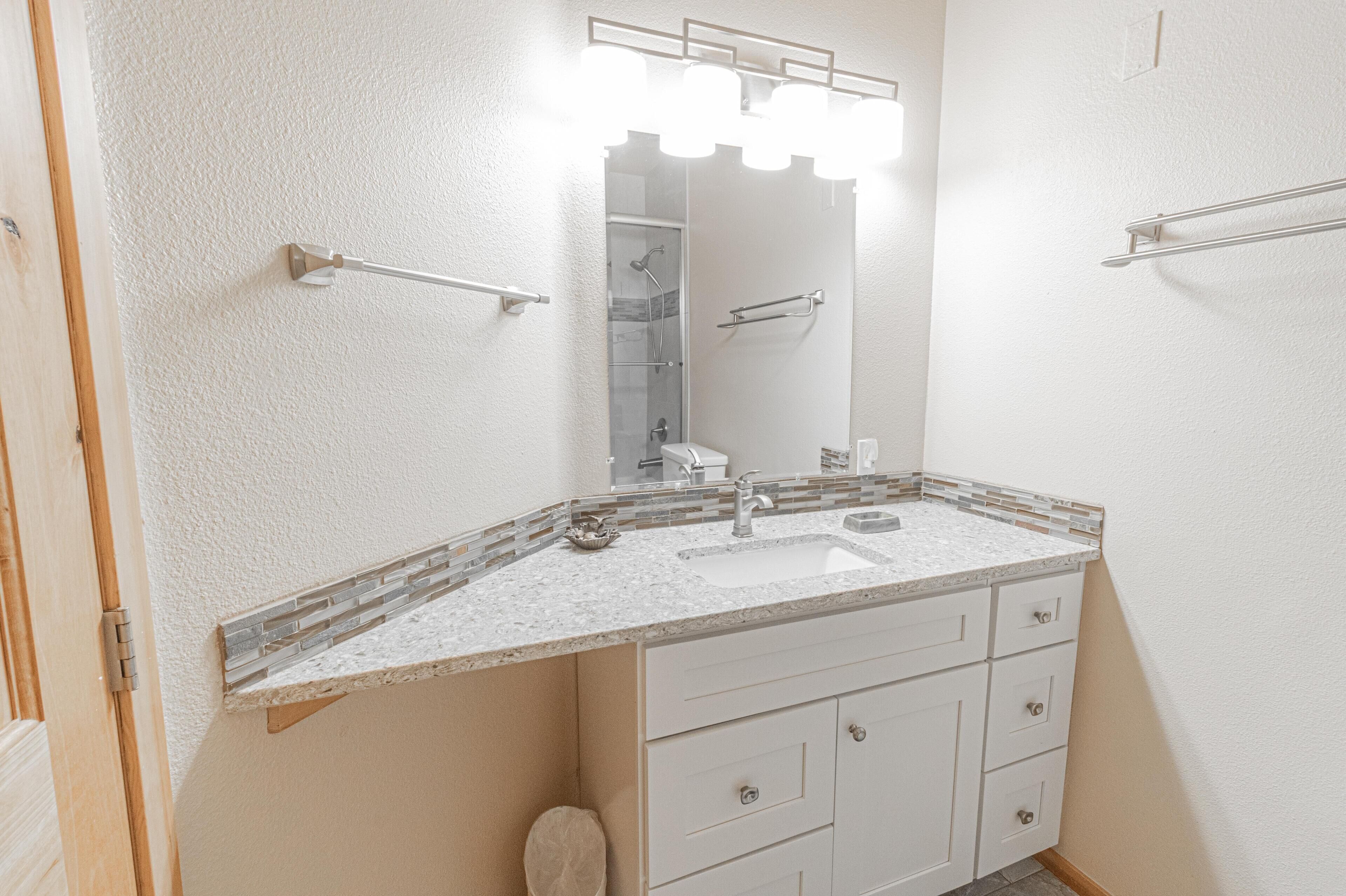A bathroom with a white sink, a mirror with lights, and a shower visible in the background.