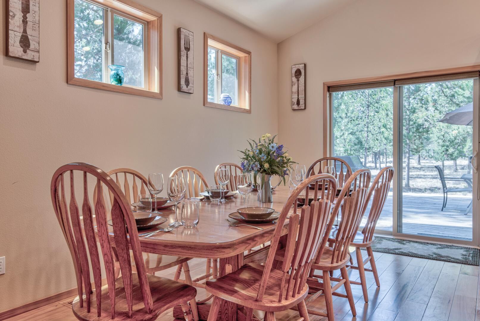 A dining area with a wooden table set for six, surrounded by six wooden chairs. There are framed artworks on the walls and a large window providing a view of the outdoors.