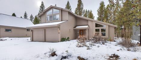 Una casa de dos pisos con garaje, rodeada de suelo y árboles cubiertos de nieve.
