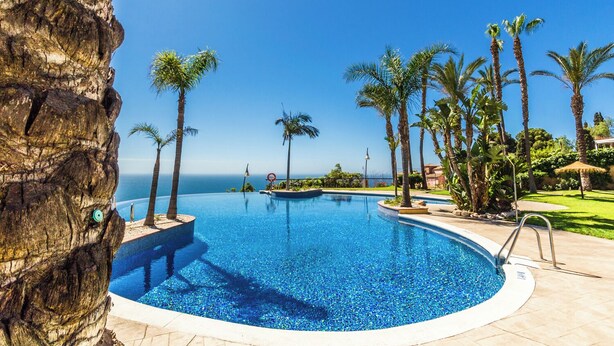 Una piscina con vista despejada al océano, rodeada de palmeras y un acantilado rocoso.