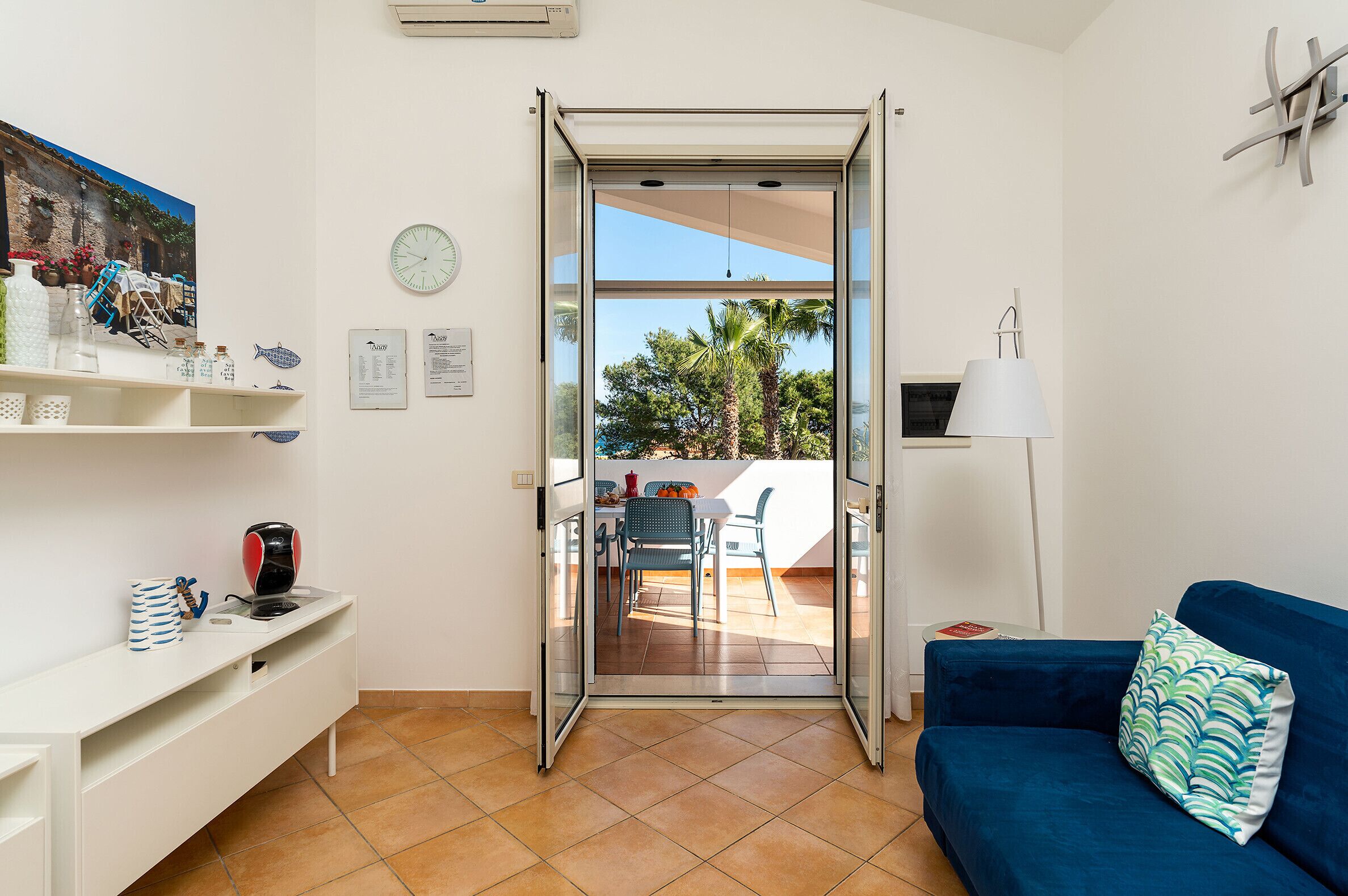 A room with a sliding glass door leading to an outdoor patio, a blue sofa, a white desk with shelves, and a clock on the wall.