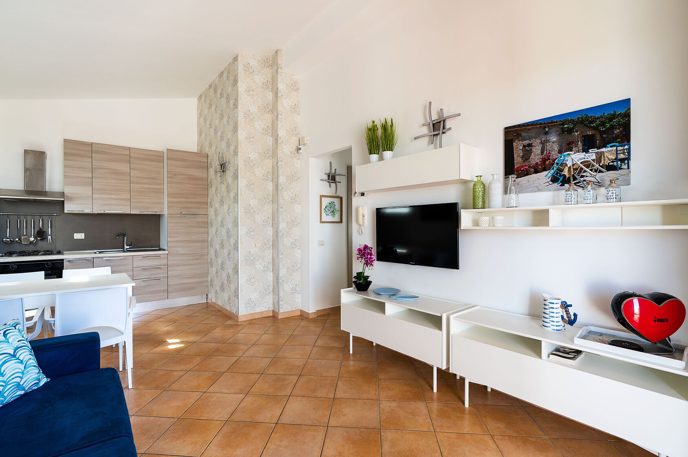 A modern kitchen with white cabinets, a dining area with a blue sofa, and a living room with a flat-screen TV and white entertainment unit.