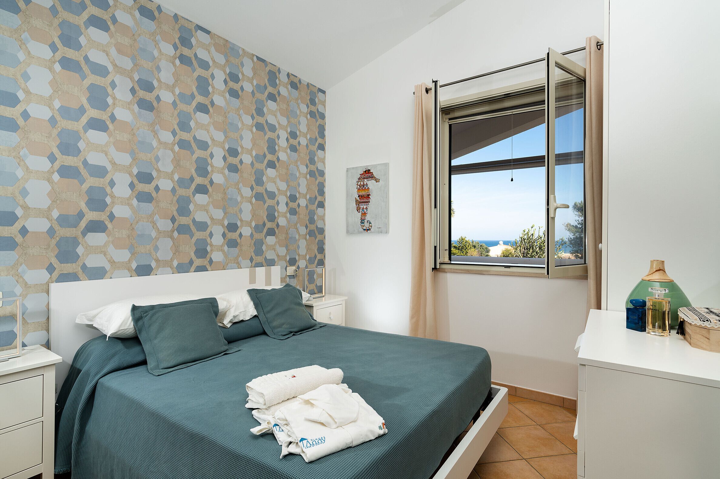 Un lit bien fait, recouvert d’une couvre-lit verte, une table de chevet avec une lampe, une fenêtre donnant sur un beau paysage et un mur orné d’un motif géométrique.