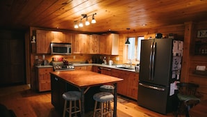A wooden cabin-style kitchen with a central island, bar stools, stainless steel appliances, and a refrigerator. - Log Home in a Peaceful Wood in Jim Thorpe (Jim Thorpe)