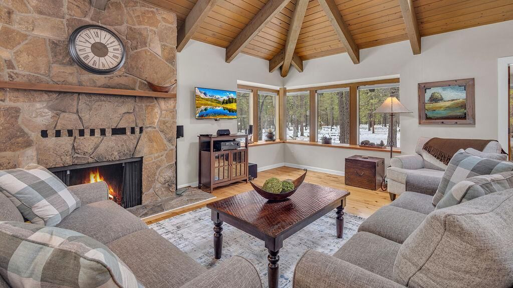Un salon chaleureux avec une cheminée en pierre, un plafond en bois et de grandes fenêtres donnant sur le paysage enneigé.