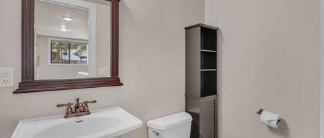 Un baño con un lavabo blanco, un inodoro, un espejo con marco de madera y una estantería.