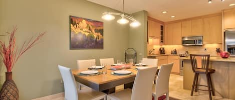 A dining area with a table set for four, a kitchen with wooden cabinets, and a painting on the wall.
