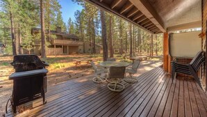 A covered deck with a grill, outdoor seating, and a view of a cabin in a wooded area. - Awbrey Lane #7 by Village Properties at Sunriver (Bend)