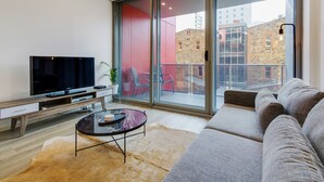 Uma sala de estar moderna com uma TV de tela plana, uma mesa de centro de vidro, um sofá e uma porta de correr de vidro que dá para uma área externa com uma parede vermelha e vista para edifícios.