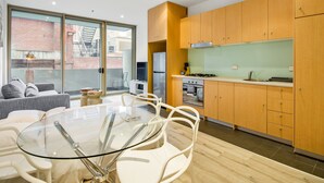 Cozinha moderna com armários de madeira, mesa de jantar de vidro e vista para um edifício de tijolos.