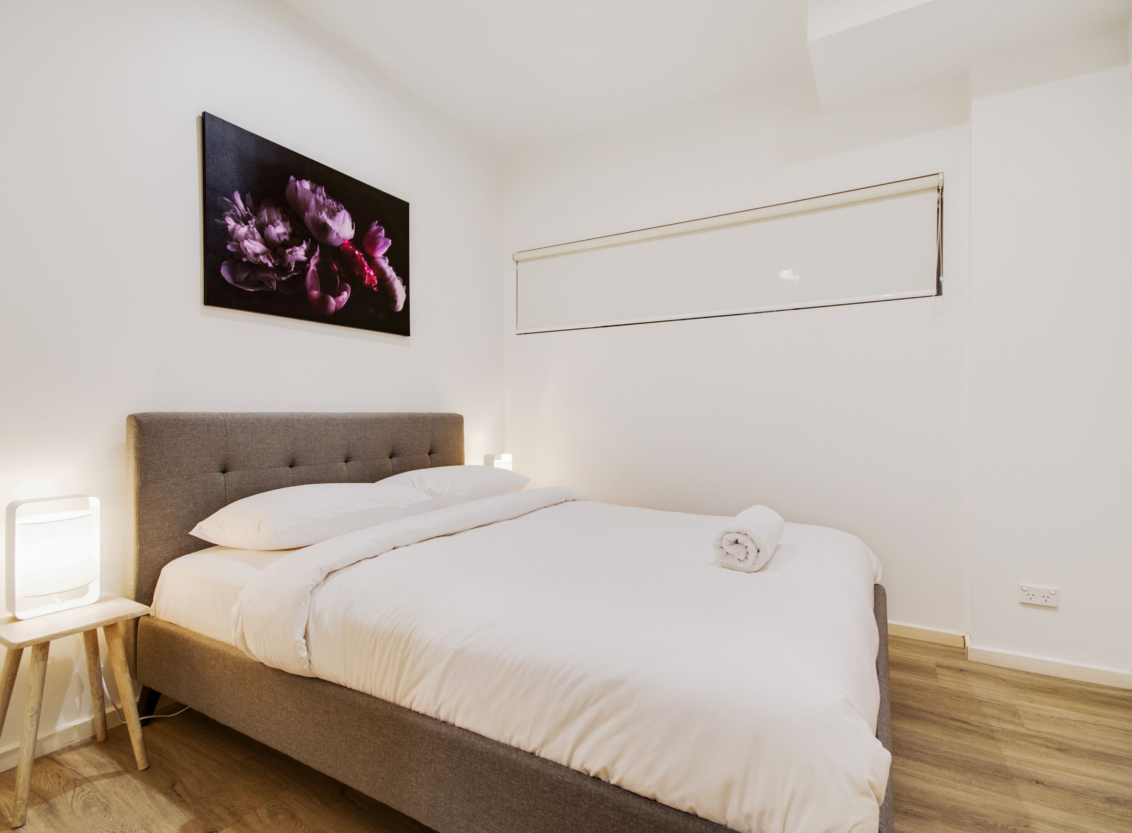 Un letto rifatto con lenzuola bianche, una testiera grigia imbottita e un comodino con una lampada.