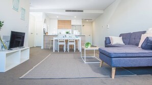 Una sala de estar moderna con un sofá gris, un televisor sobre un mueble blanco y una zona de comedor con una mesa y sillas blancas.