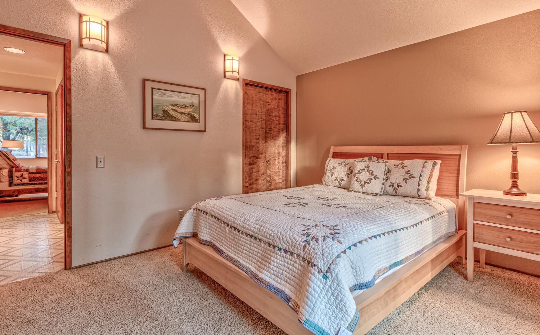 A bedroom with a large bed, wooden headboard, and a nightstand with a lamp. There is a framed picture on the wall and a door leading to another room.