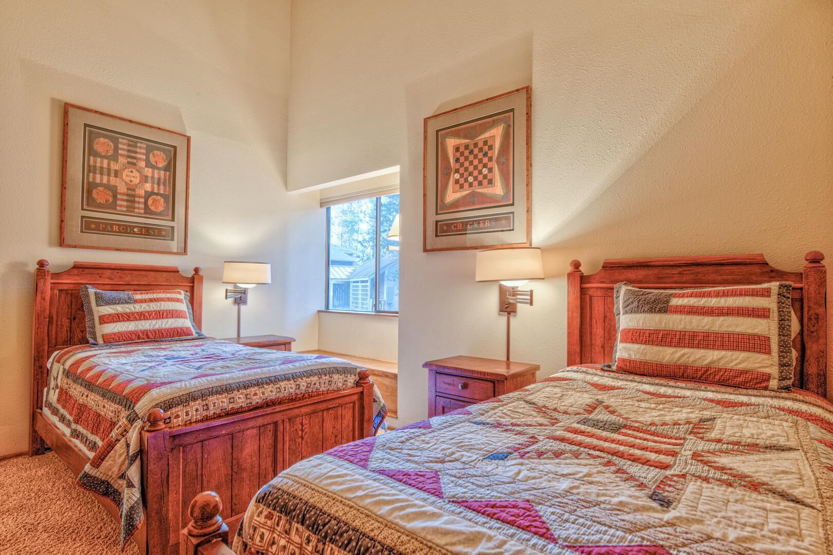 A bedroom with two wooden beds, each with a quilted bedspread and a pillow with an American flag design. There are two bedside tables with lamps, and a window providing natural light.