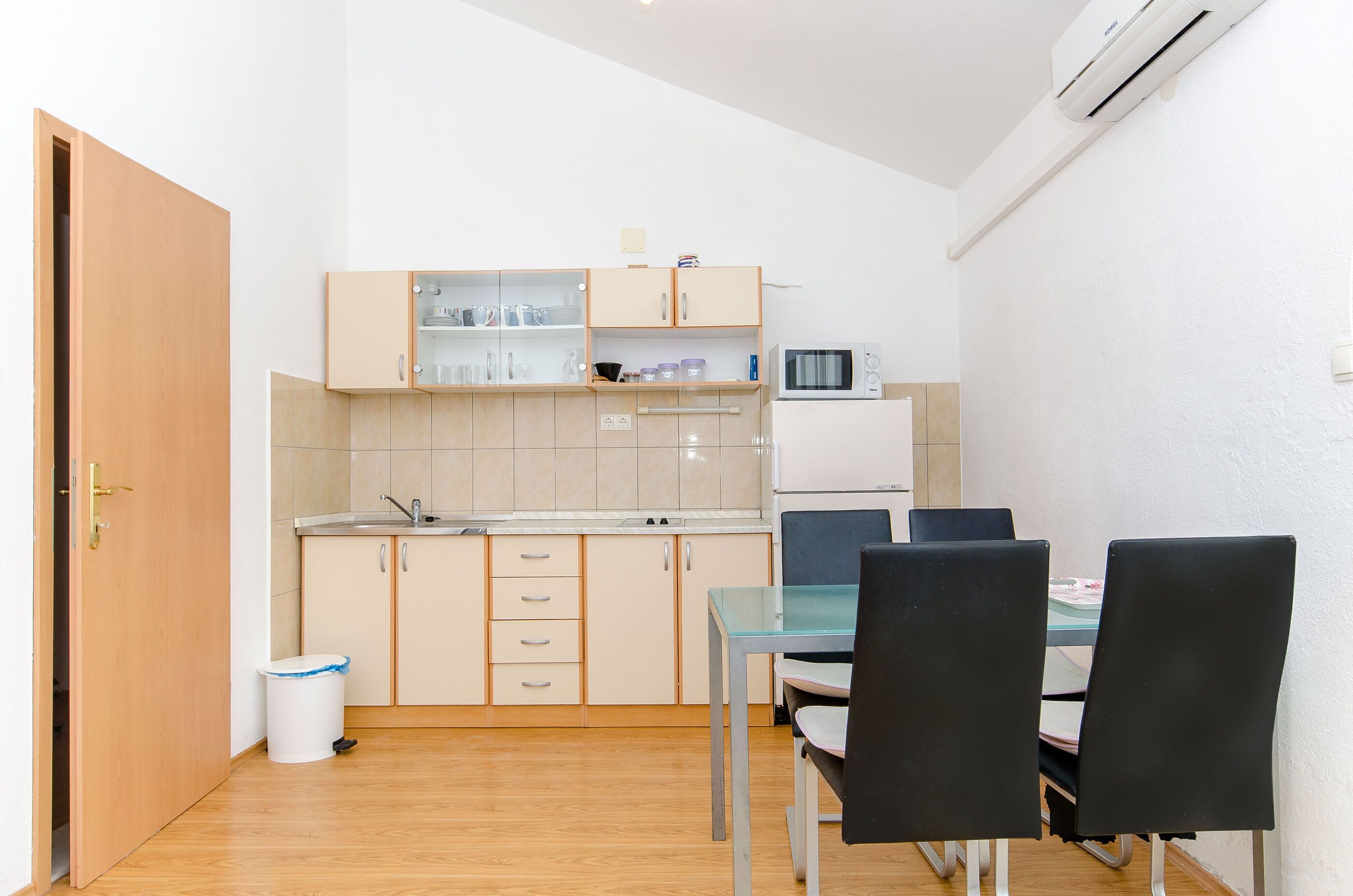 A compact kitchen with a microwave, sink, and cabinets. There is a dining table with chairs and a trash bin.