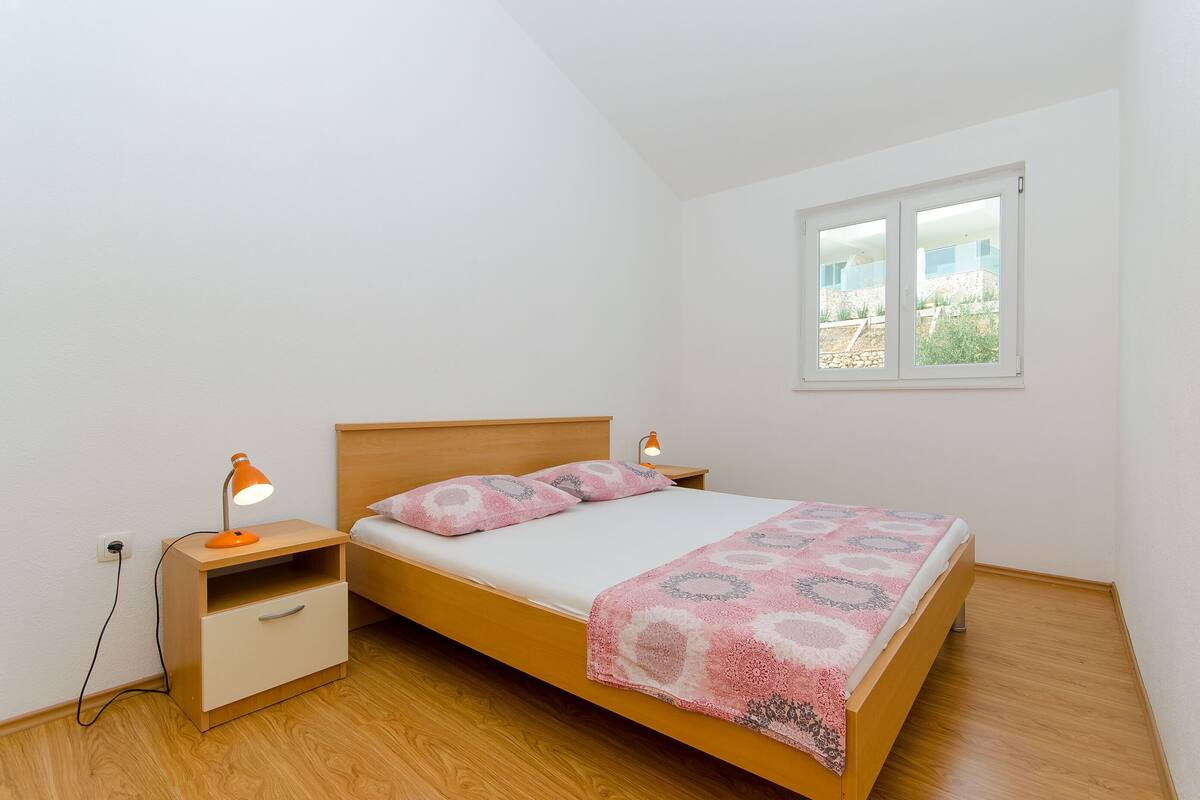 A single bed with a wooden headboard, a bedside table with a lamp, and a window with a view of greenery.