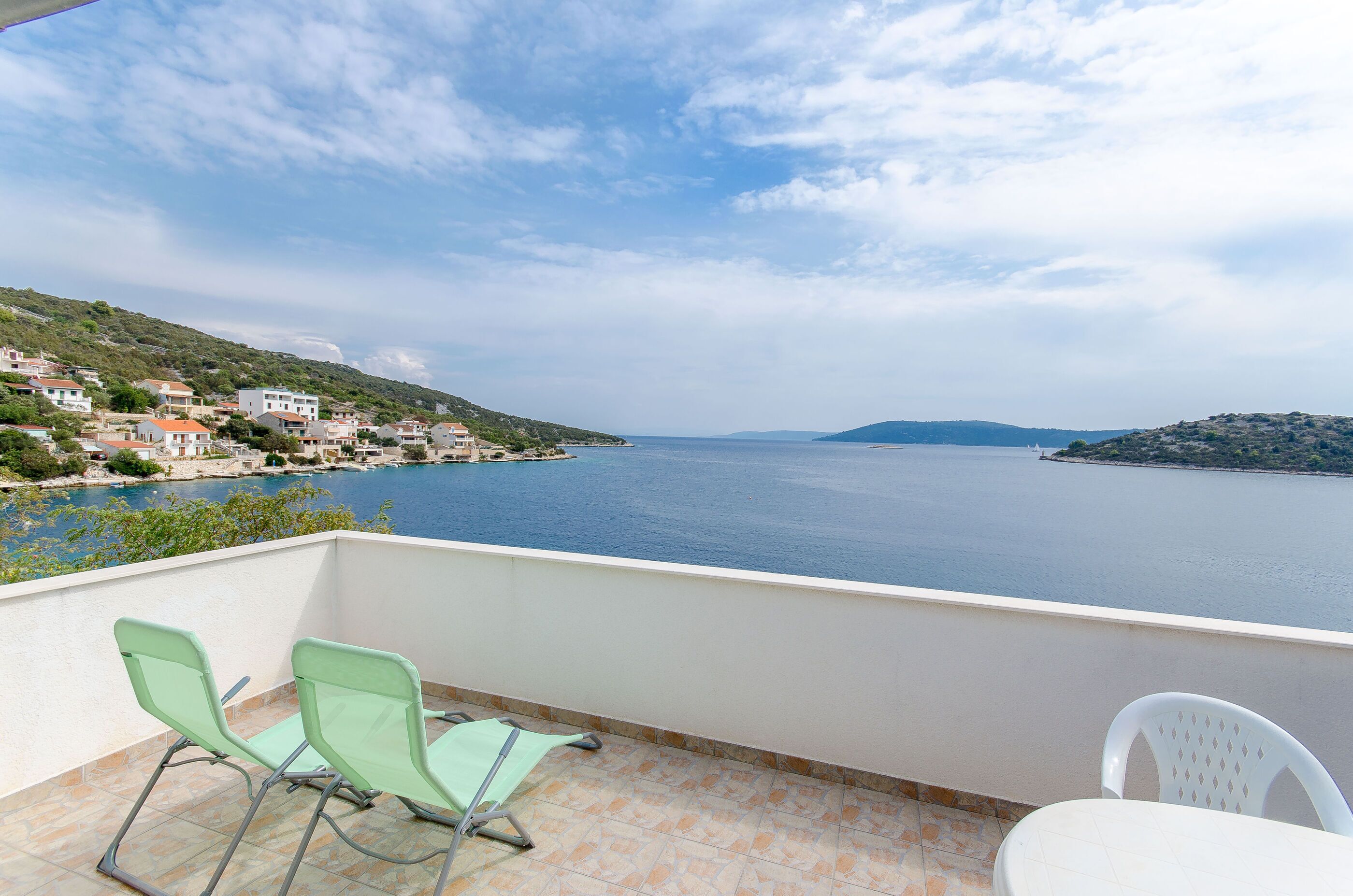 A balcony with two green chairs and a white table, overlooking a calm sea and a coastal village.